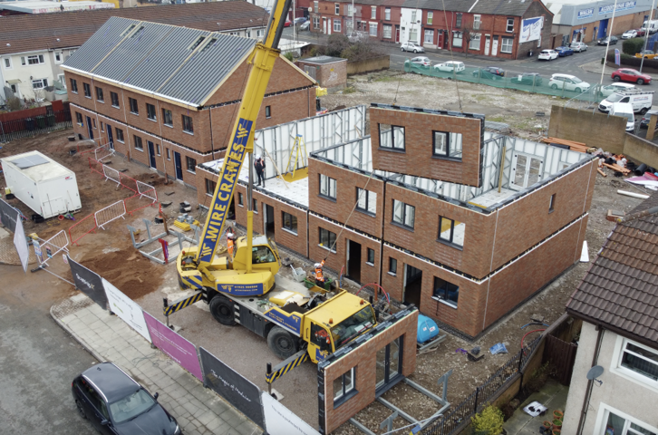 A crane lifts a modular wall section into place as new brick homes are built on a residential street.