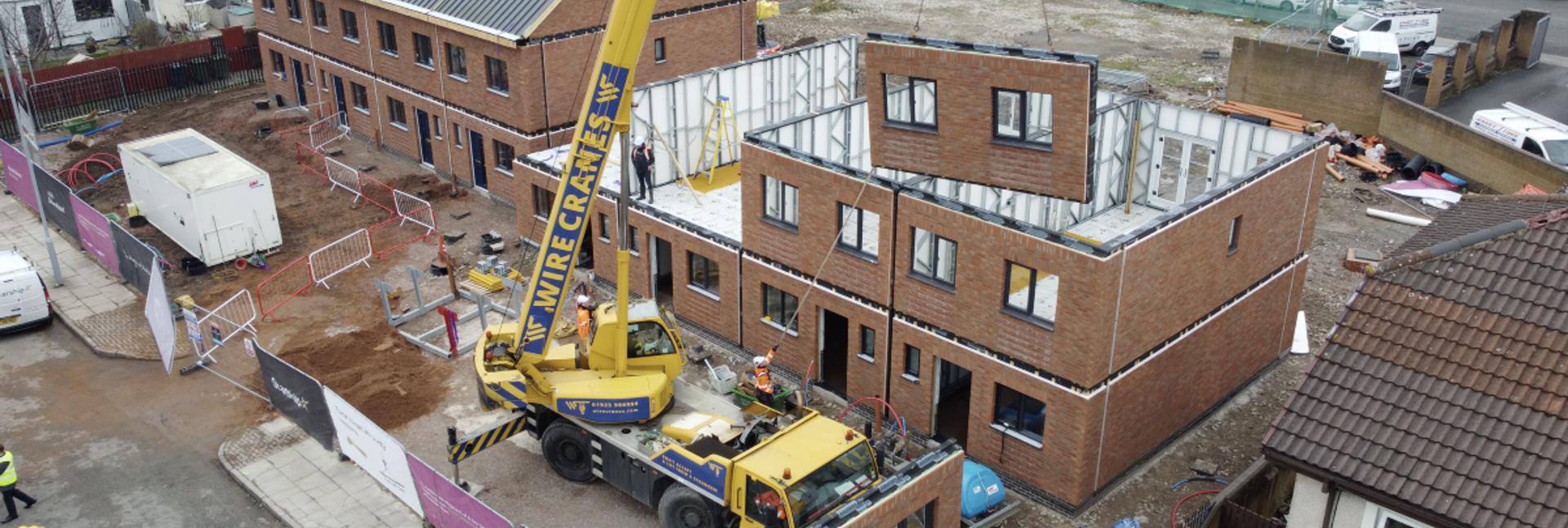 A crane lifts a modular wall section into place as new brick homes are built on a residential street.