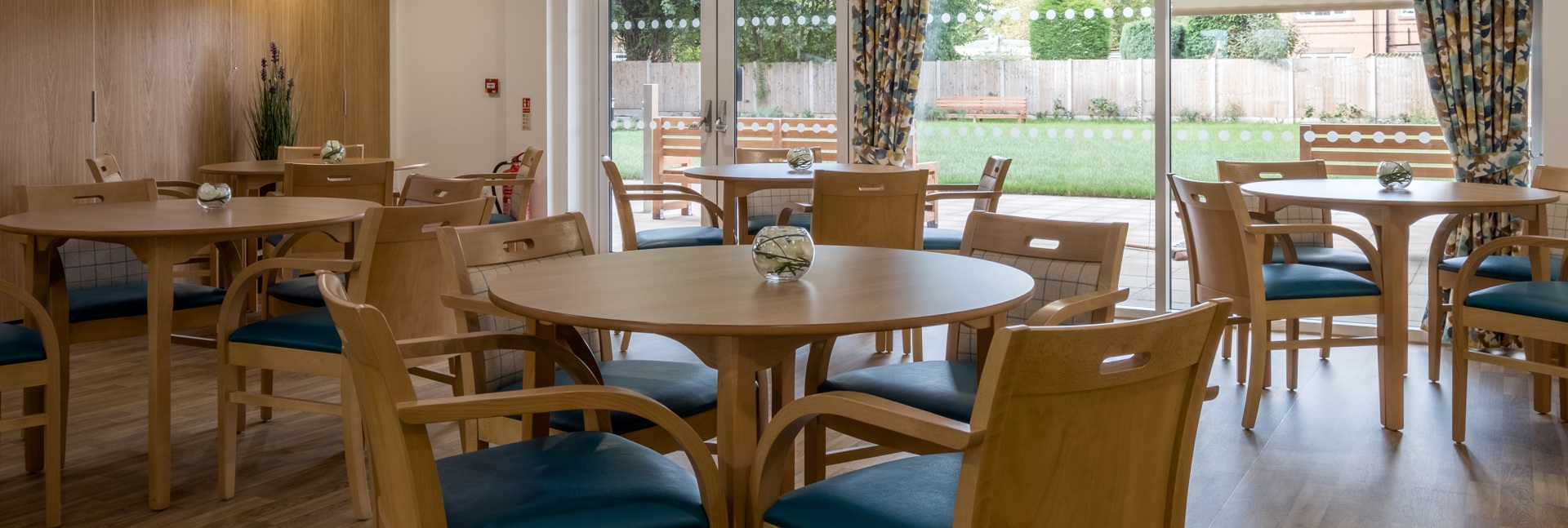 A bright communal dining room with wooden tables and chairs overlooking a garden through large windows.