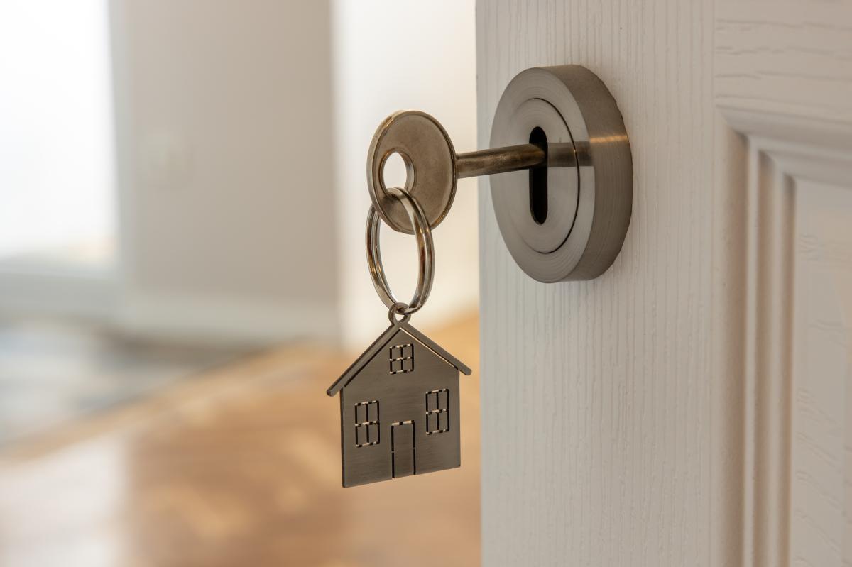 A close‑up of a key in a door, with a house‑shaped keyring hanging from it.