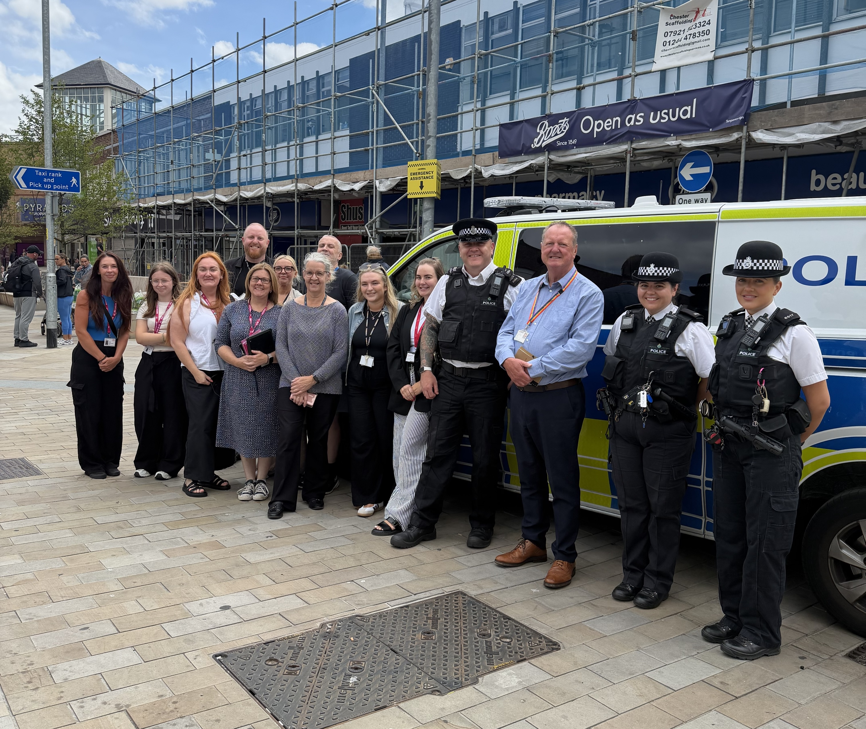 Group photo of Magenta Living colleagues, police officers and partner organisations standing beside a marked police van at a community anti-social behaviour (ASB) awareness event in a town centre. The group is gathered outdoors on a pedestrianised street, with shops visible in the background.