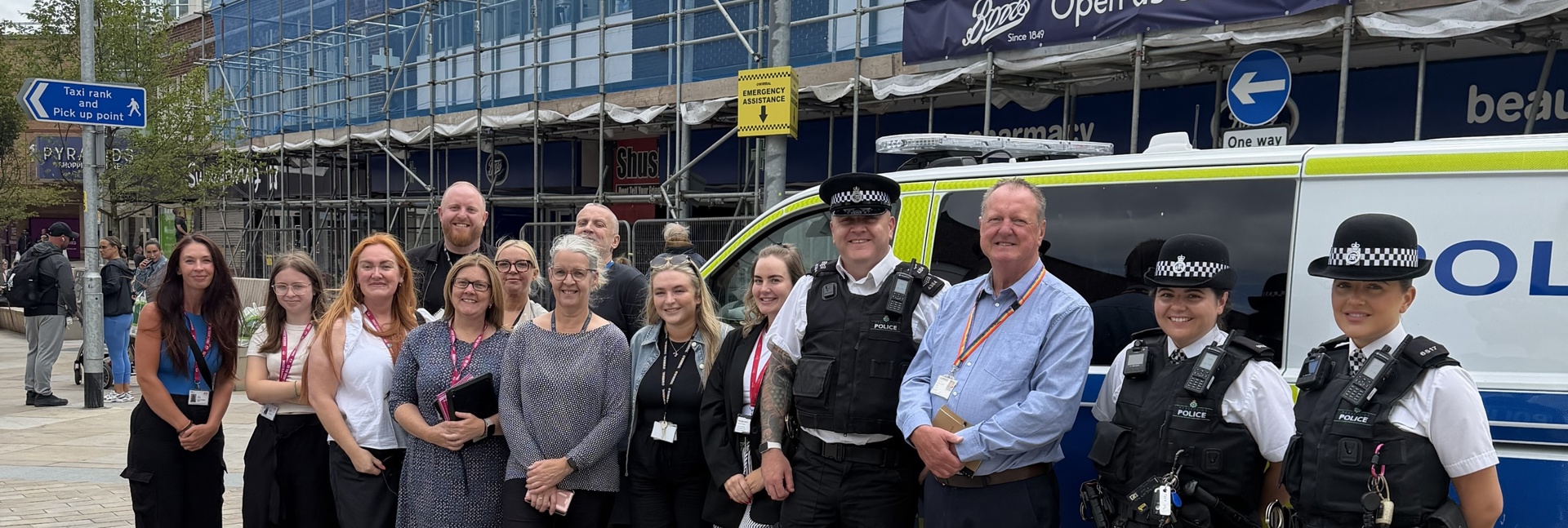 Group photo of Magenta Living colleagues, police officers and partner organisations standing beside a marked police van at a community anti-social behaviour (ASB) awareness event in a town centre. The group is gathered outdoors on a pedestrianised street, with shops visible in the background.