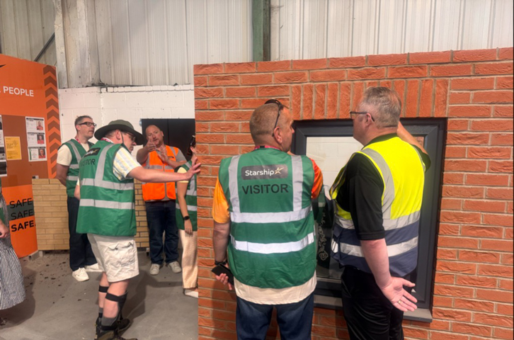 Members of the Magenta Customer and Communities Committee tour Starship's Birkenhead factory, where visitors in high-visibility vests examine a brick-faced wall section and window frame. The visit formed part of a development day exploring how modern construction methods are helping deliver sustainable, carbon zero homes for local communities.