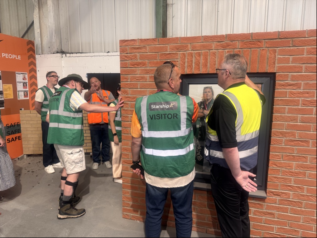 Members of the Magenta Customer and Communities Committee tour Starship's Birkenhead factory, where visitors in high-visibility vests examine a brick-faced wall section and window frame. The visit formed part of a development day exploring how modern construction methods are helping deliver sustainable, carbon zero homes for local communities.