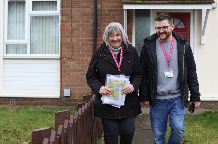Two colleagues walking along a path outside homes, carrying documents and wearing ID lanyards.