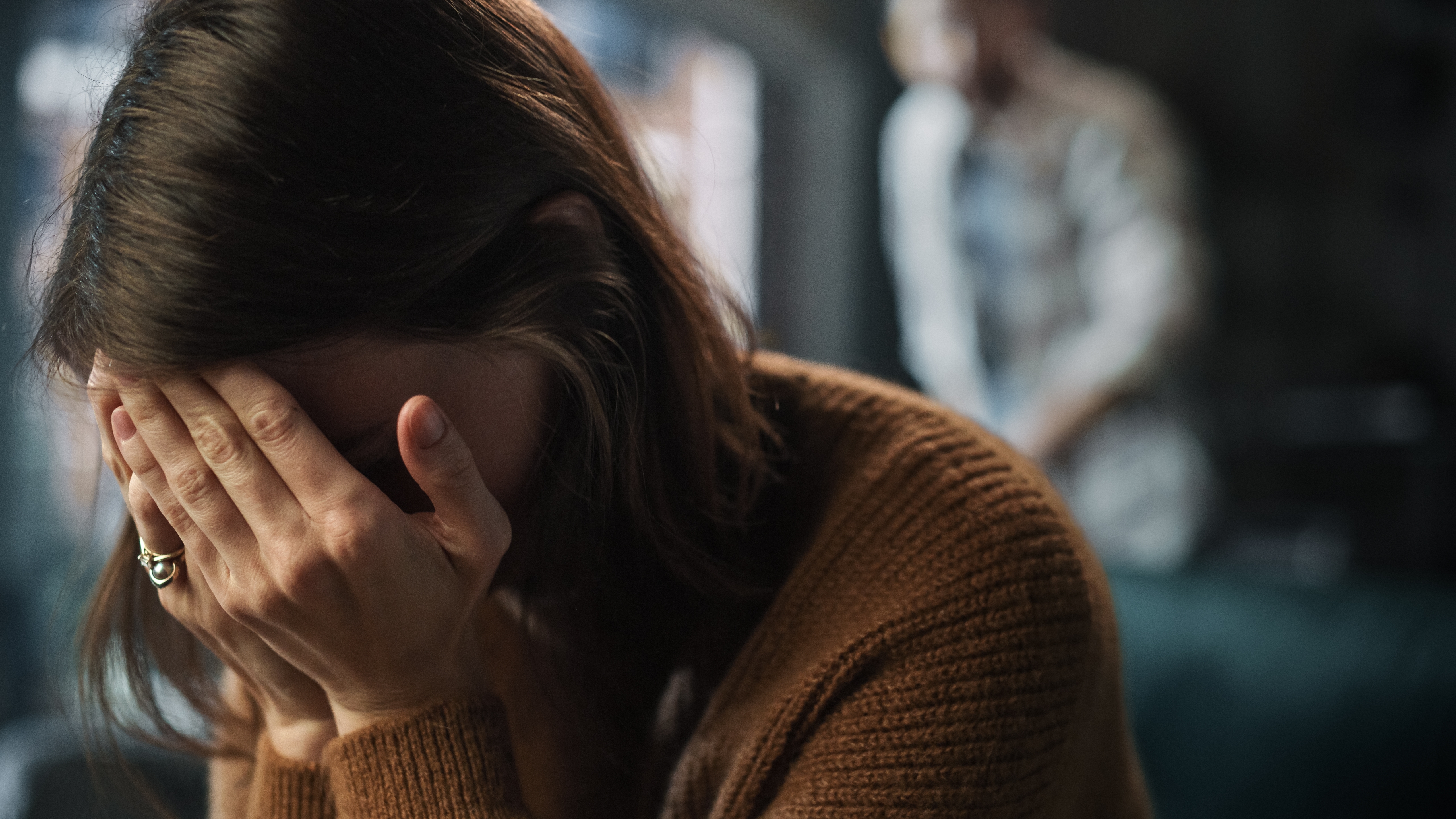 A person sitting with their head in their hands while someone stands in the background.