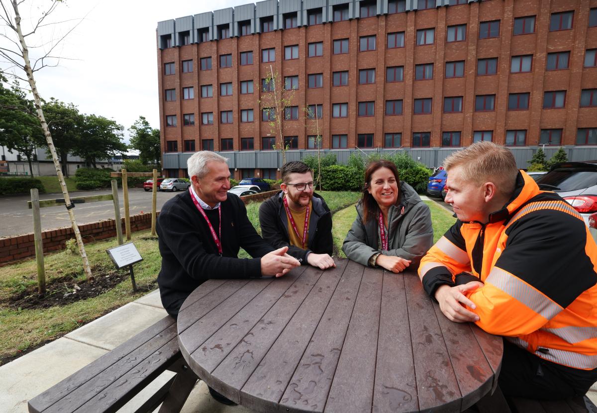Four people sitting at an outdoor picnic table having a discussion outside a large brick building.