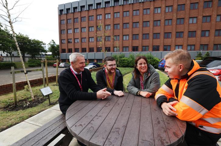 Four people sitting at an outdoor picnic table having a discussion outside a large brick building.