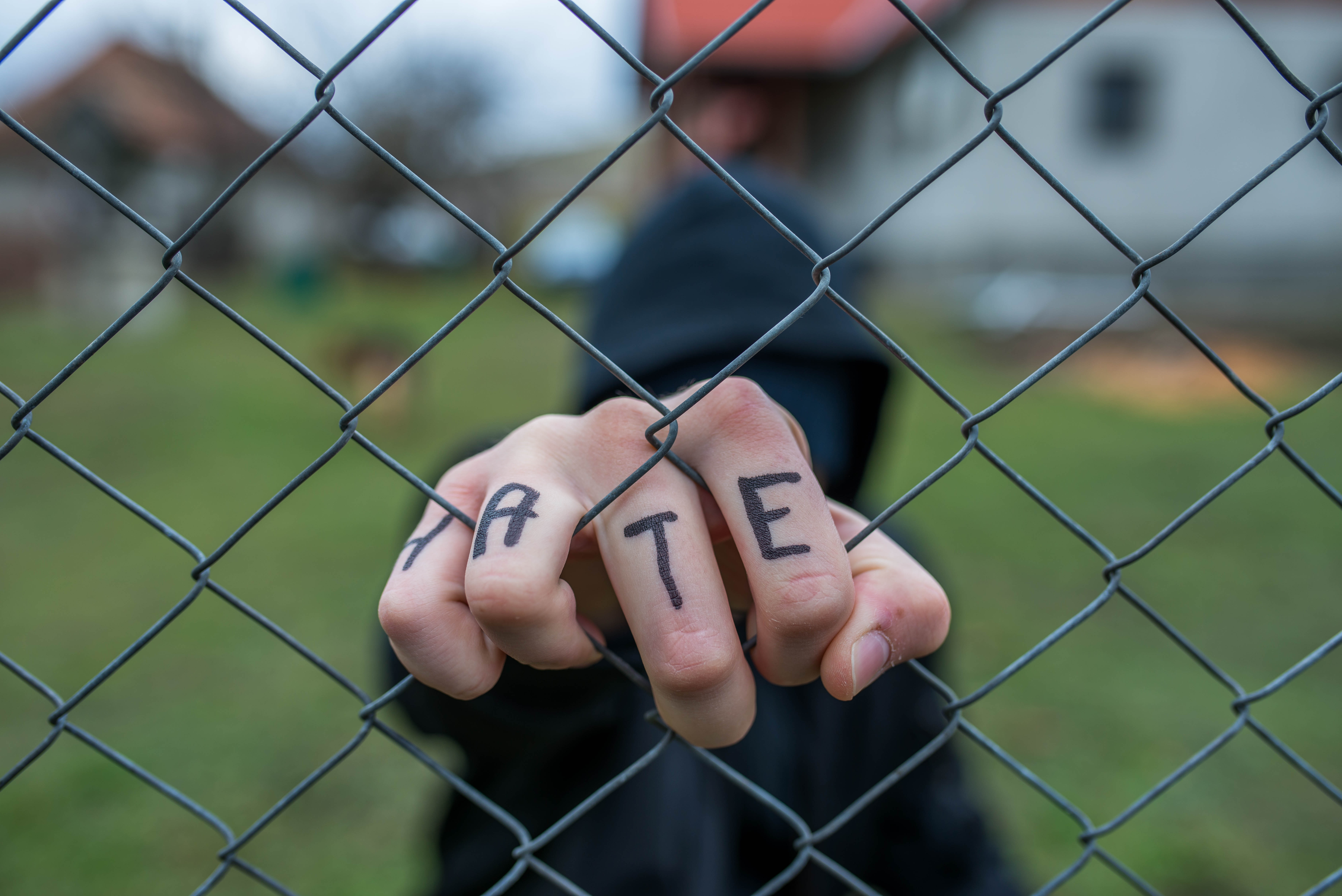A person in a hooded top holding their fist against a chain‑link fence, with ‘HATE’ written on their knuckles.
