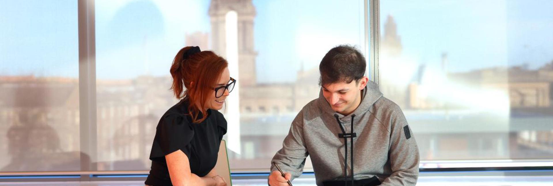 A conversation taking place at a desk, with one person writing notes and large windows showing buildings in the background.