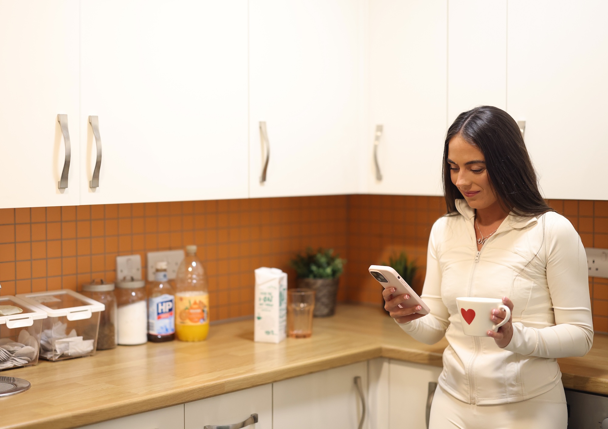 A person standing in a kitchen holding a mug and looking at a mobile phone.