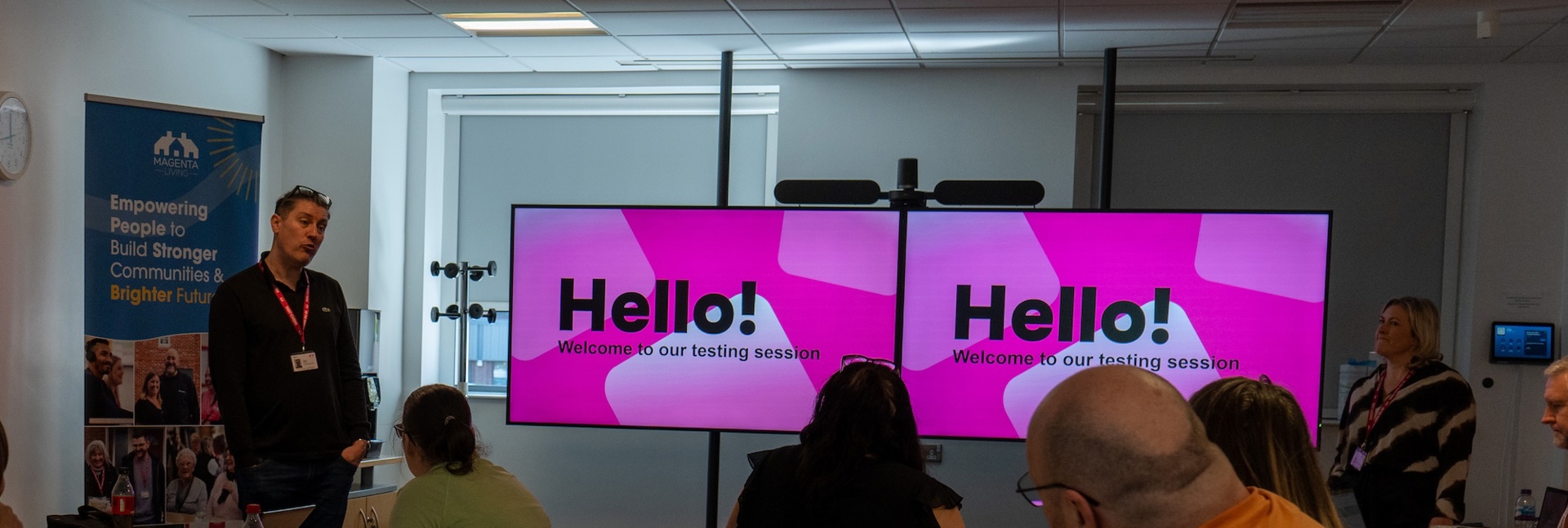 People seated in a meeting room watch a presentation on two screens reading “Hello! Welcome to our testing session.”