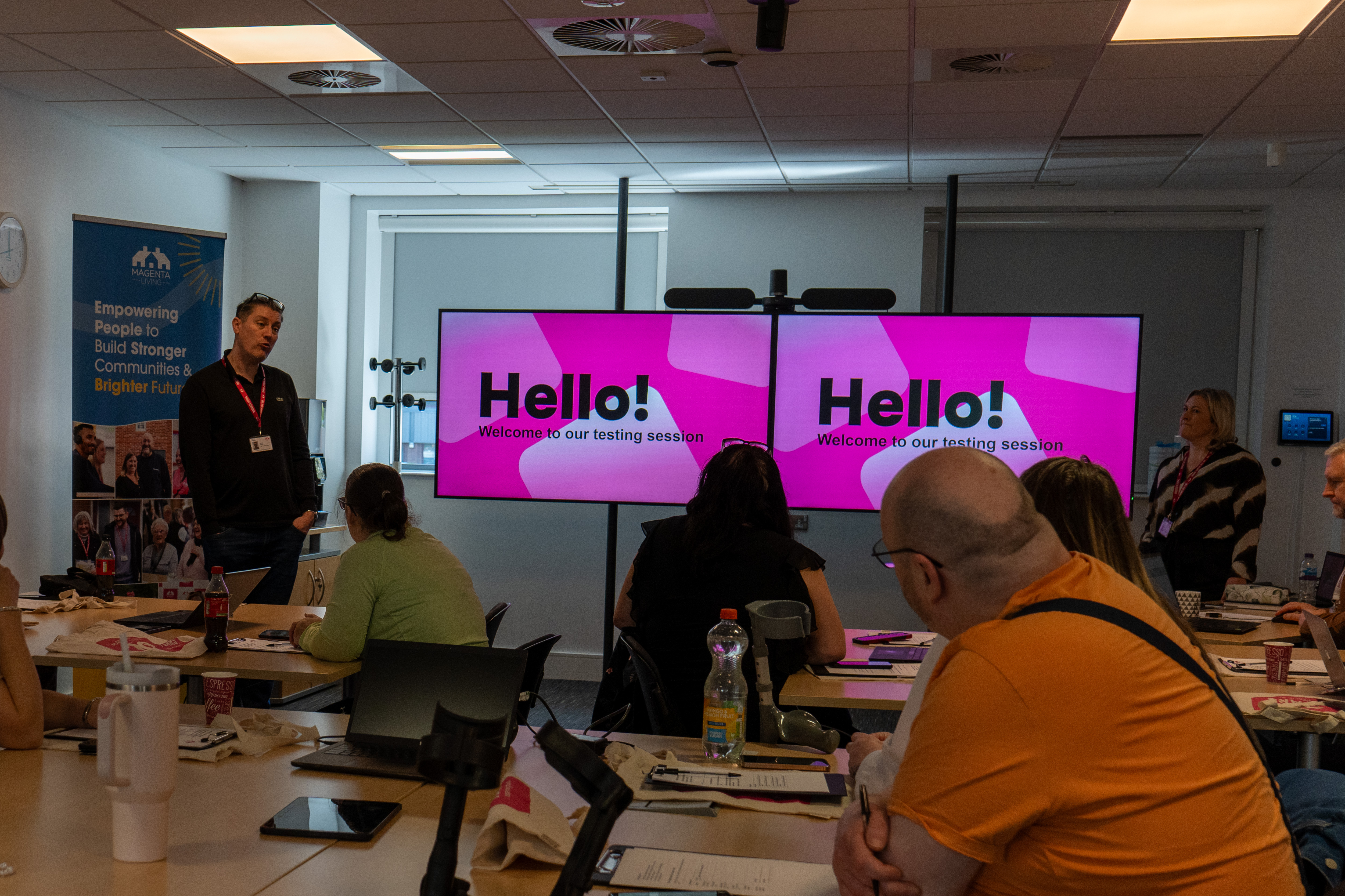 People seated in a meeting room watch a presentation on two screens reading “Hello! Welcome to our testing session.”