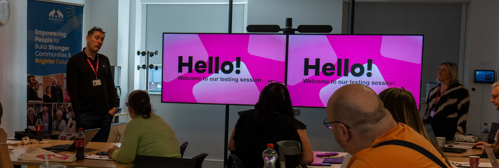 People seated in a meeting room watch a presentation on two screens reading “Hello! Welcome to our testing session.”