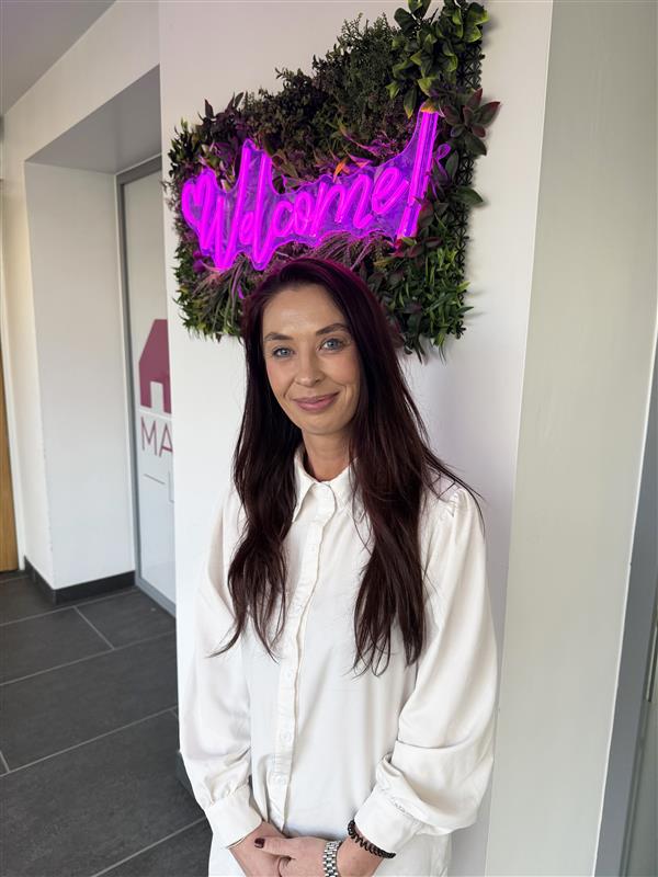 A person standing in a reception area beneath a bright neon “Welcome!” sign framed with greenery.