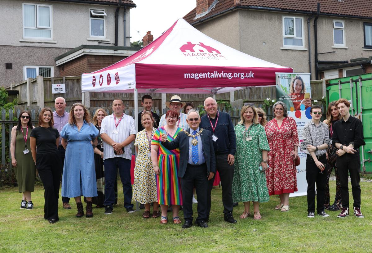 Colleagues and community members gathered on a lawn beneath a Magenta Living-branded tent during an outdoor event.