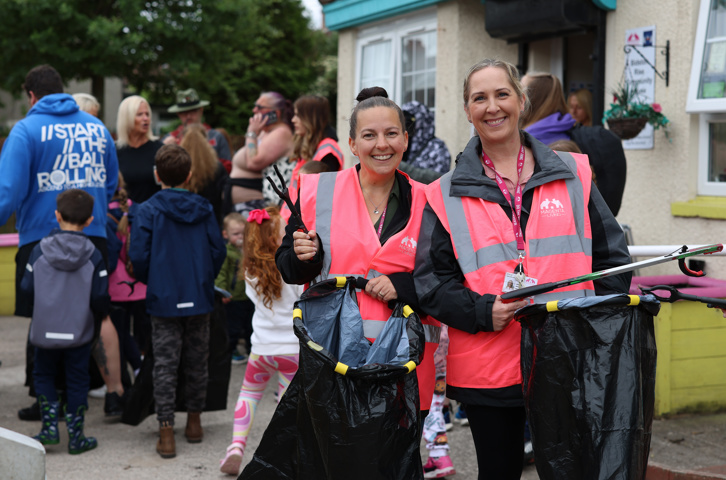 Two women in pink high‑visibility vests smile while holding litter‑picking tools, with people gathered behind them on a residential street.