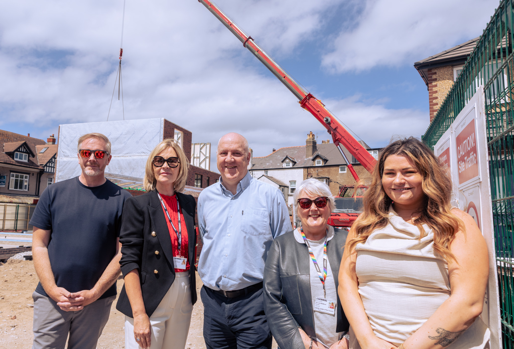 Five people stand together on a construction site in front of a partially built structure, while a red crane lifts materials overhead.
