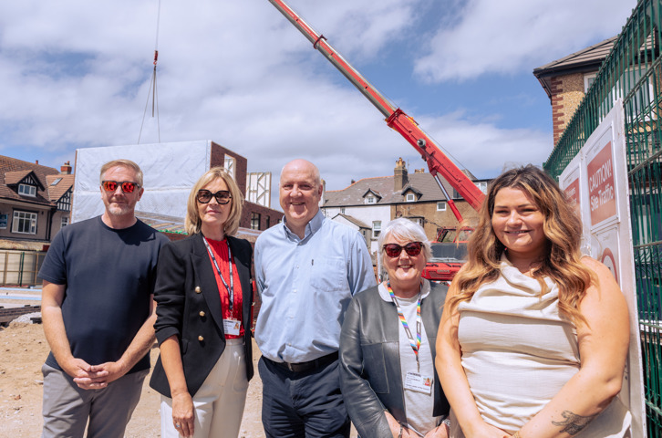 Five people stand together on a construction site in front of a partially built structure, while a red crane lifts materials overhead.