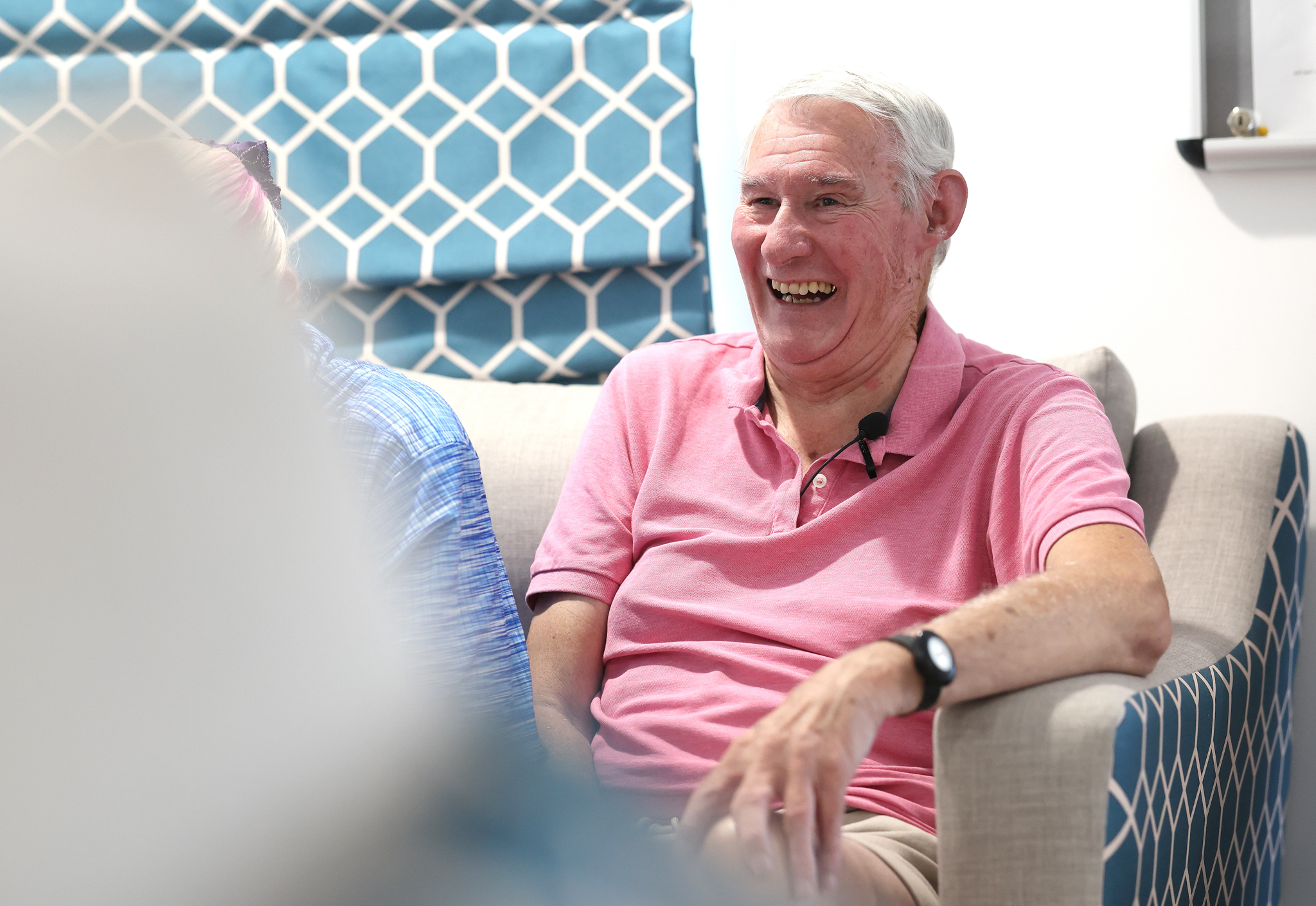 Person wearing a pink polo shirt seated in an armchair during a group discussion in a community setting.