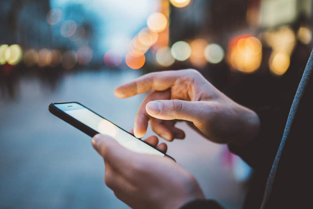 Hands holding a smartphone and tapping the screen on a busy street at dusk.