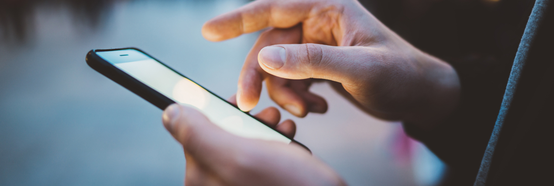 Hands holding a smartphone and tapping the screen on a busy street at dusk.