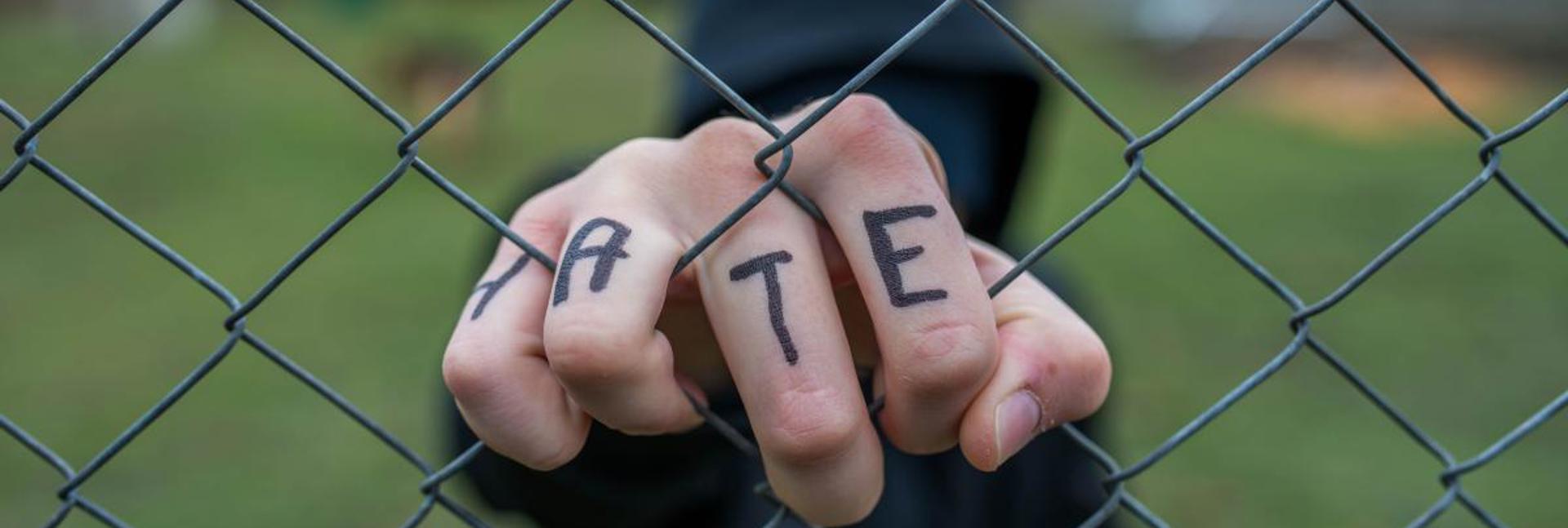 A person in a hooded jacket gripping a wire fence with the word “HATE” written across their fingers.