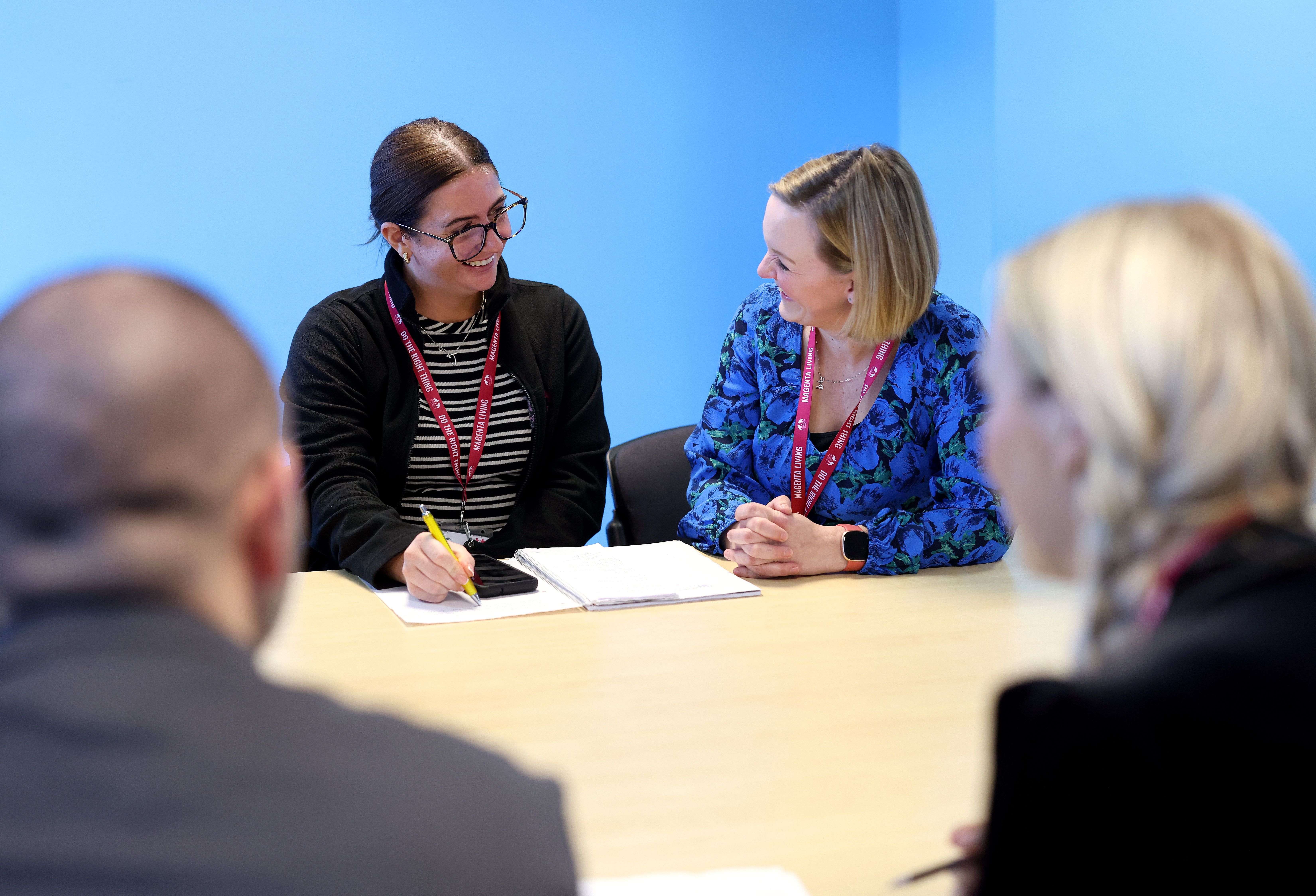 Two people sitting at a table in a meeting, writing notes and talking while others sit nearby.
