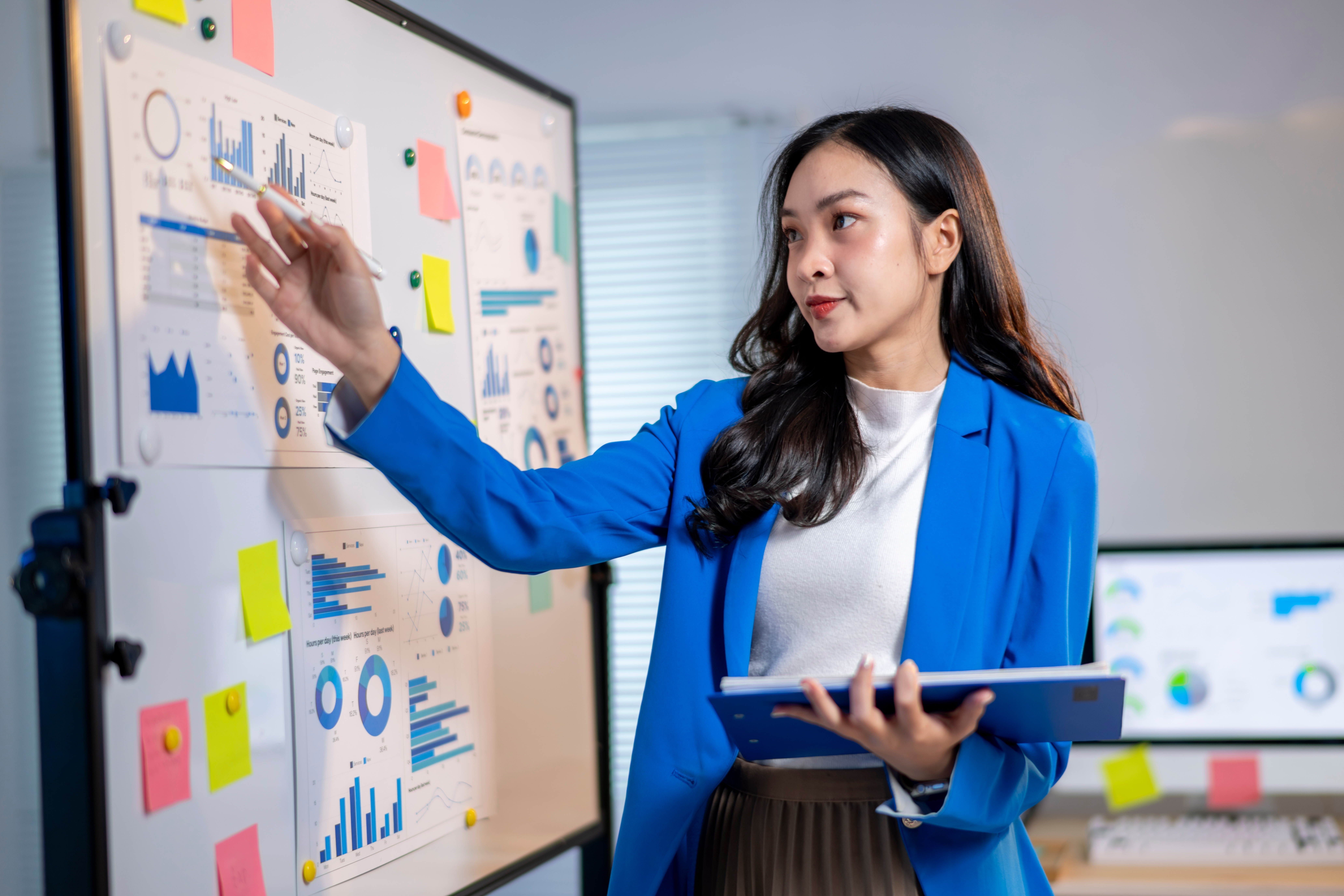 A person in a blue blazer presents data displayed on a whiteboard covered with charts, graphs and colourful sticky notes while holding a tablet in an office setting.