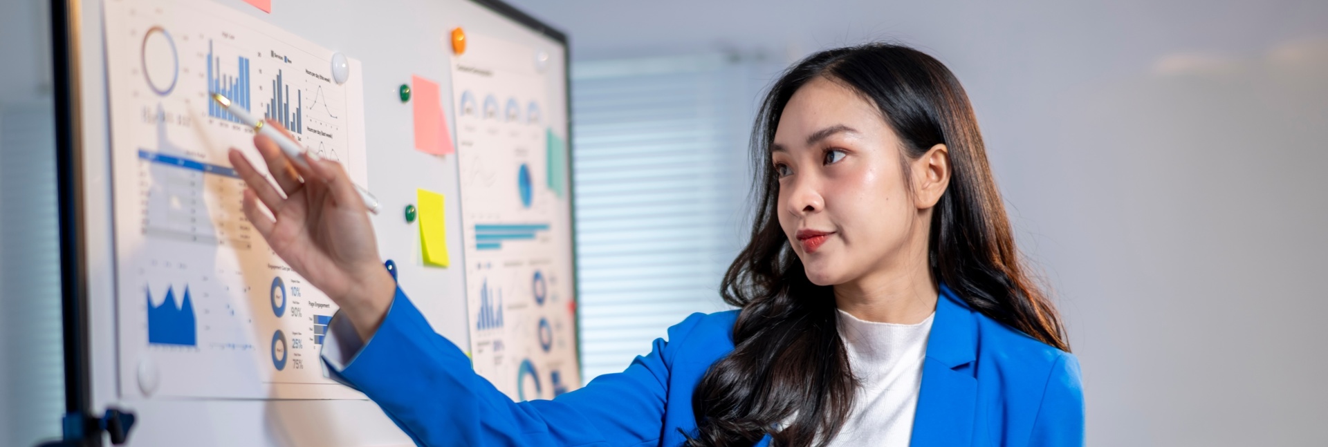 A person in a blue blazer presents data displayed on a whiteboard covered with charts, graphs and colourful sticky notes while holding a tablet in an office setting.