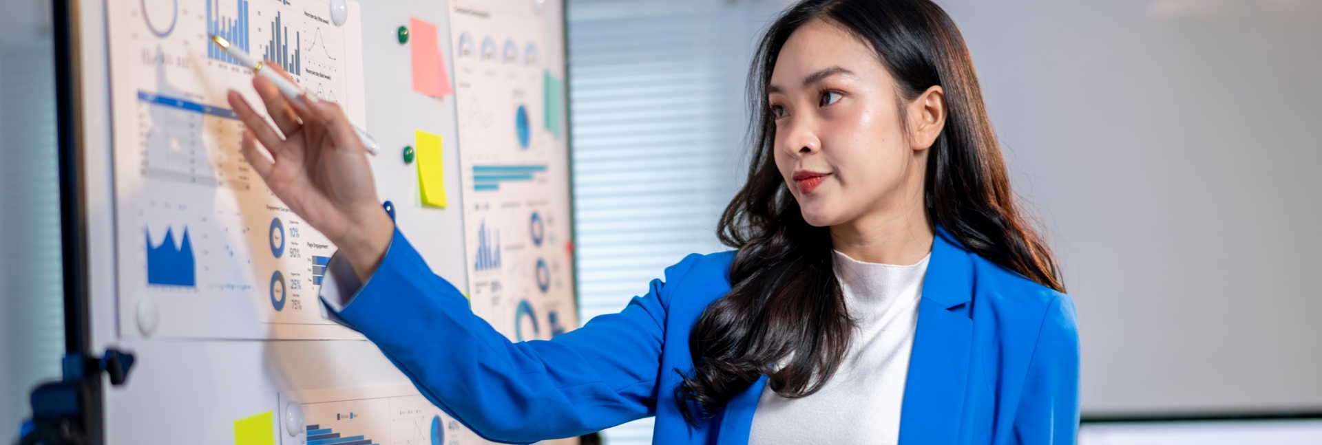 A person in a blue blazer presents data displayed on a whiteboard covered with charts, graphs and colourful sticky notes while holding a tablet in an office setting.