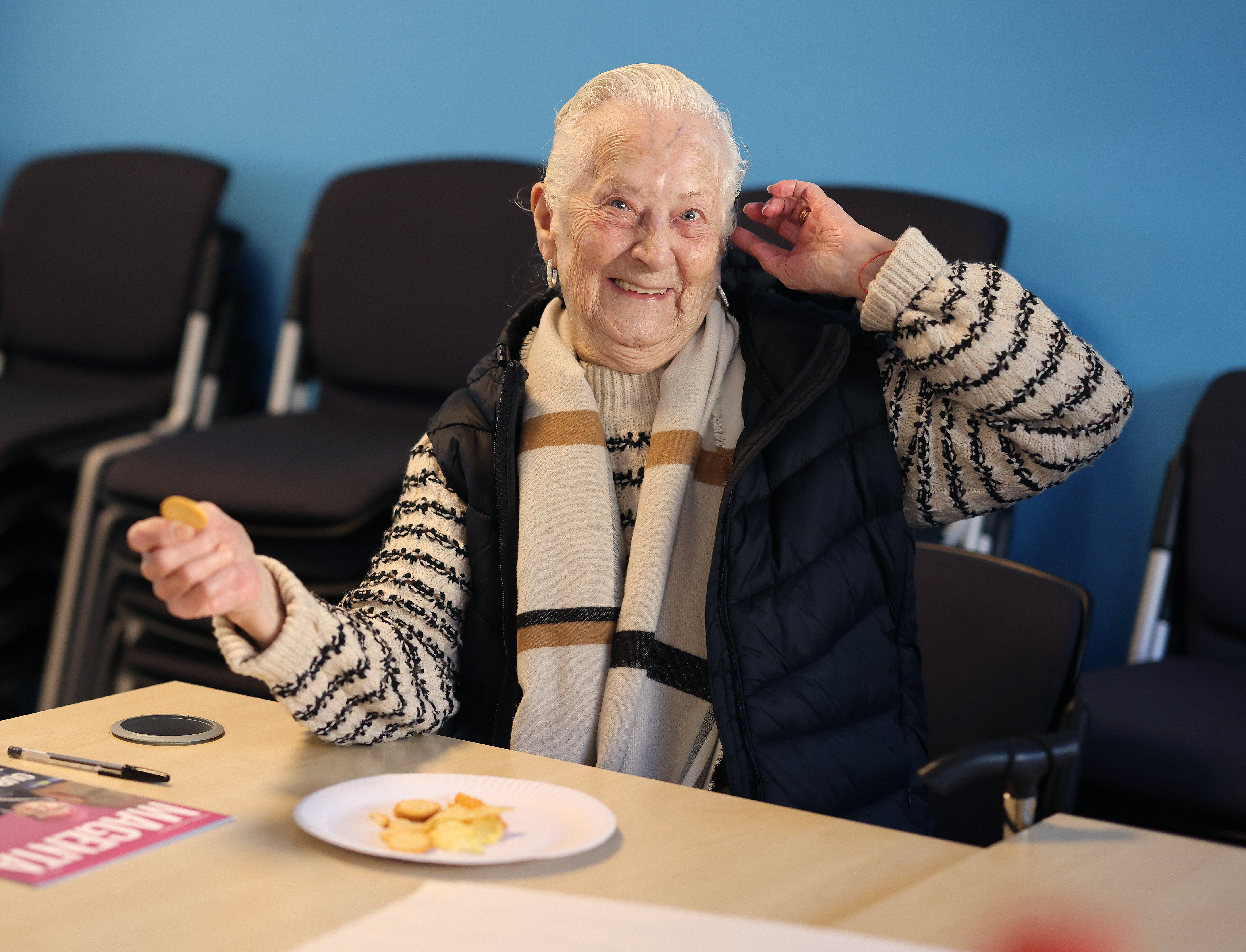 A person sitting at a table in a community room, holding a snack with a plate of food in front of them.