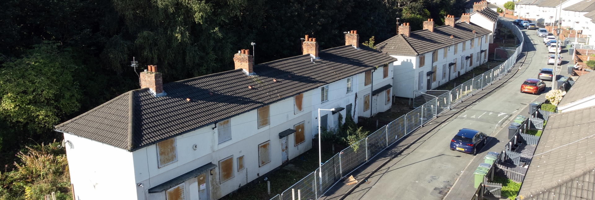 A row of empty, boarded‑up homes on a curved residential street, fenced off for safety.