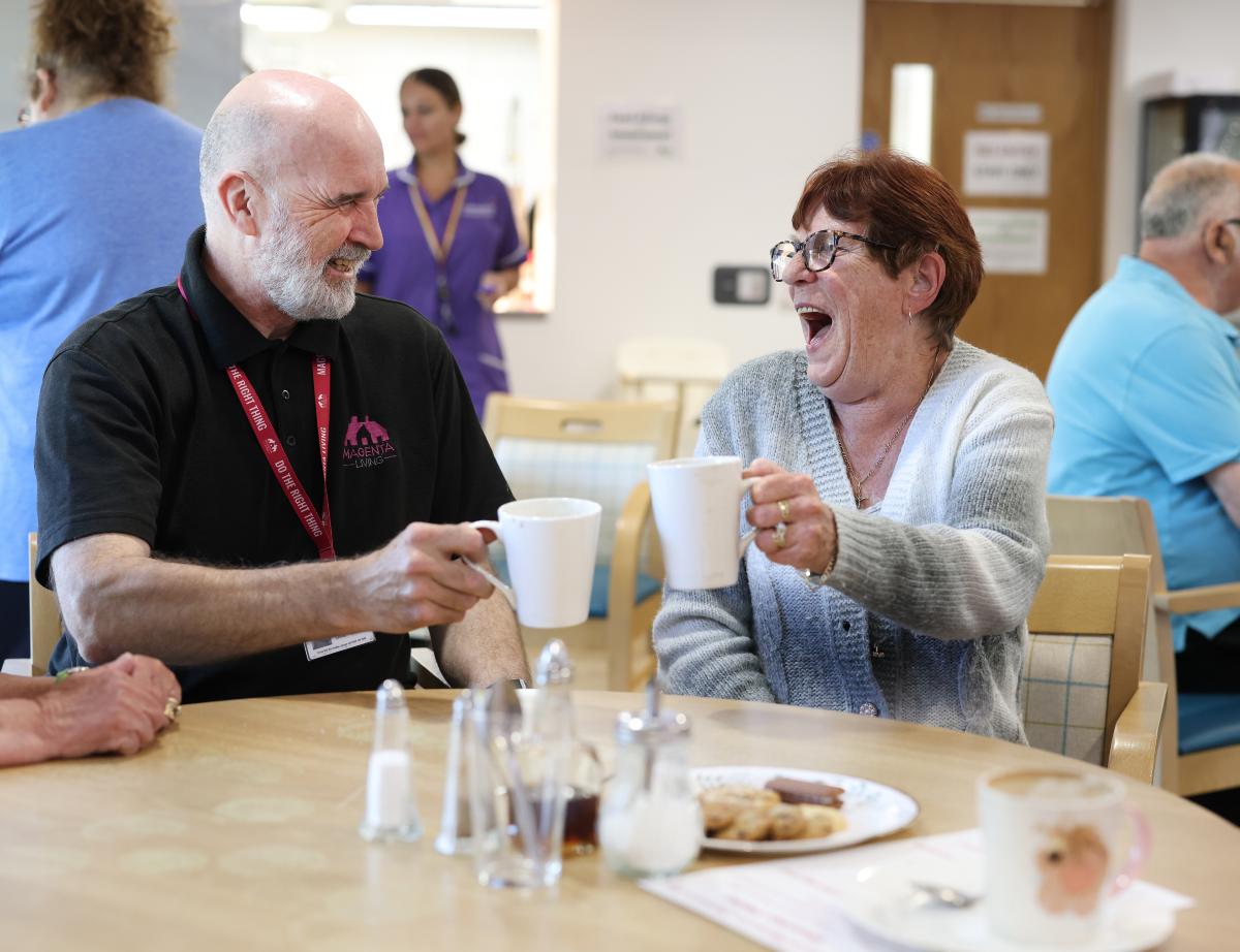 Two people sitting at a table in a community space, raising mugs during a friendly conversation.