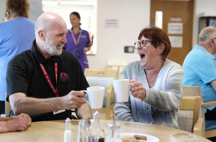 Two people sitting at a table in a community space, raising mugs during a friendly conversation.