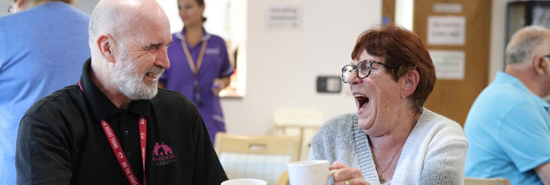 Two people sitting at a table in a community space, raising mugs during a friendly conversation.