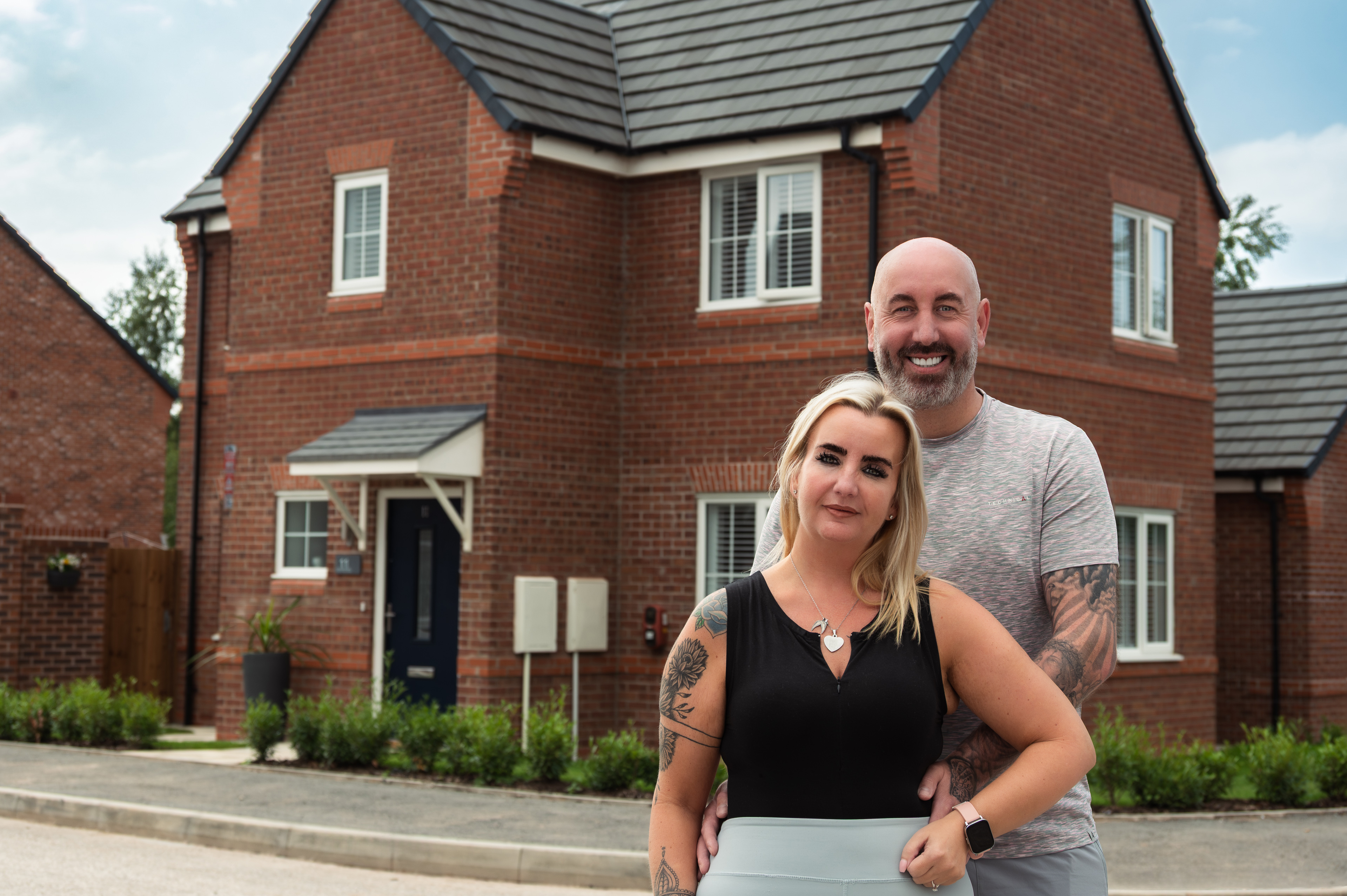 Couple standing together outside a modern red‑brick home on a residential street.