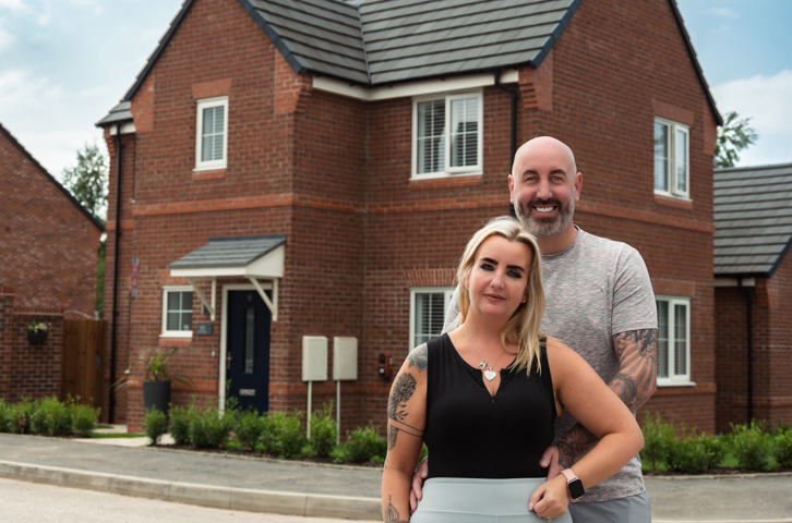 Couple standing together outside a modern red‑brick home on a residential street.