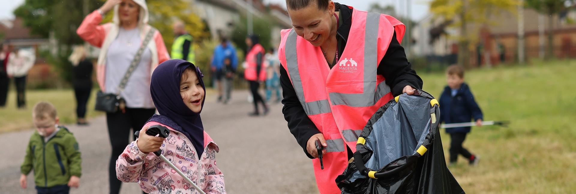 A child and an adult in high‑visibility vests taking part in a community litter‑pick, using grabbers and a bin bag.