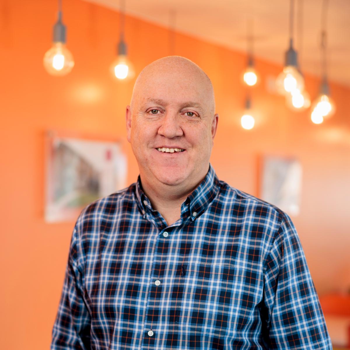 Andy Lomas wearing a checked shirt standing in a brightly lit room with hanging lightbulbs.
