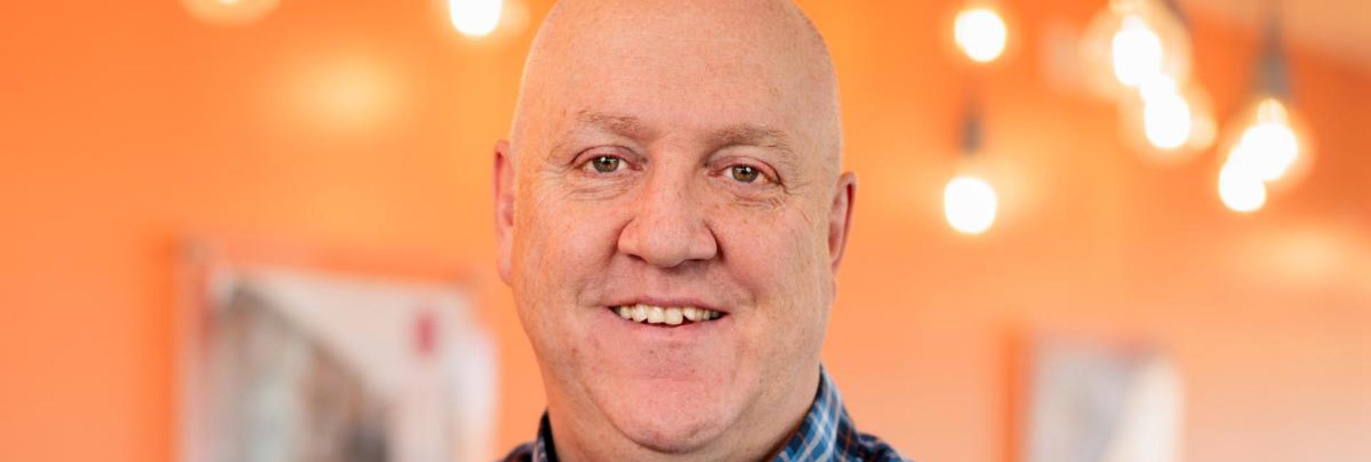 Andy Lomas wearing a checked shirt standing in a brightly lit room with hanging lightbulbs.