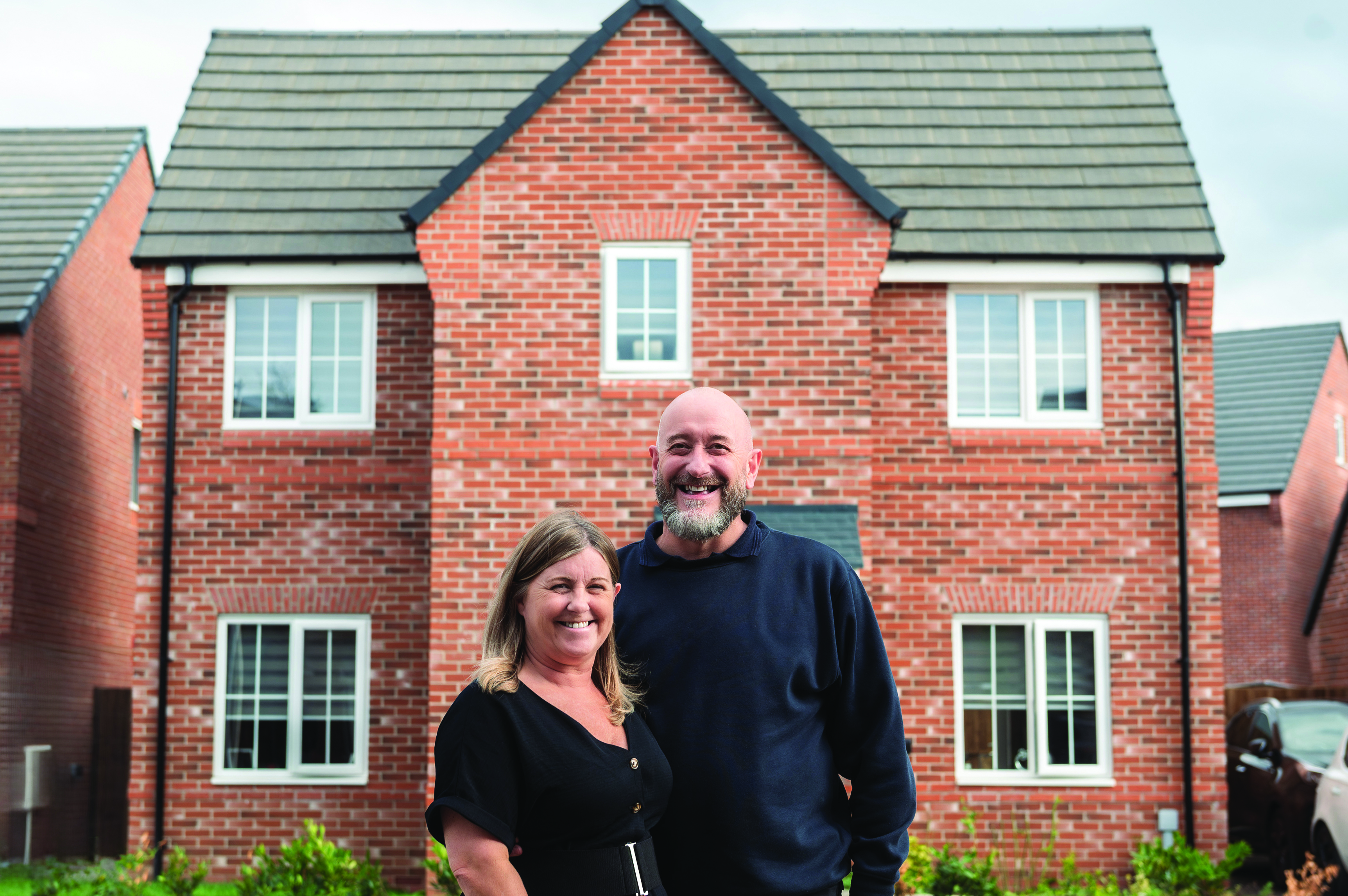 A pair of customers posing in front of a modern red‑brick house on a residential street.