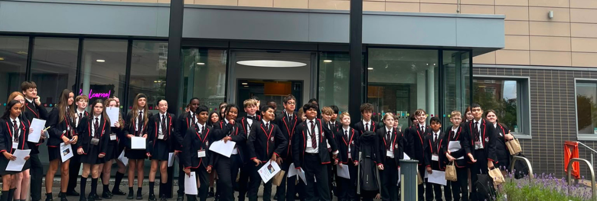 Birkenhead Park School students stand outside Magenta Living’s office building during an educational visit, holding worksheets as they stand for a group photo.