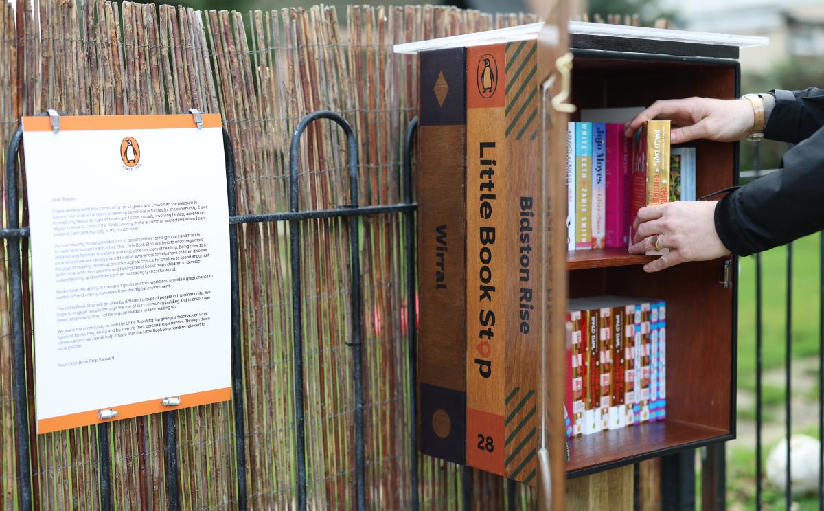 A person placing books inside a small outdoor community book stop attached to a fence.