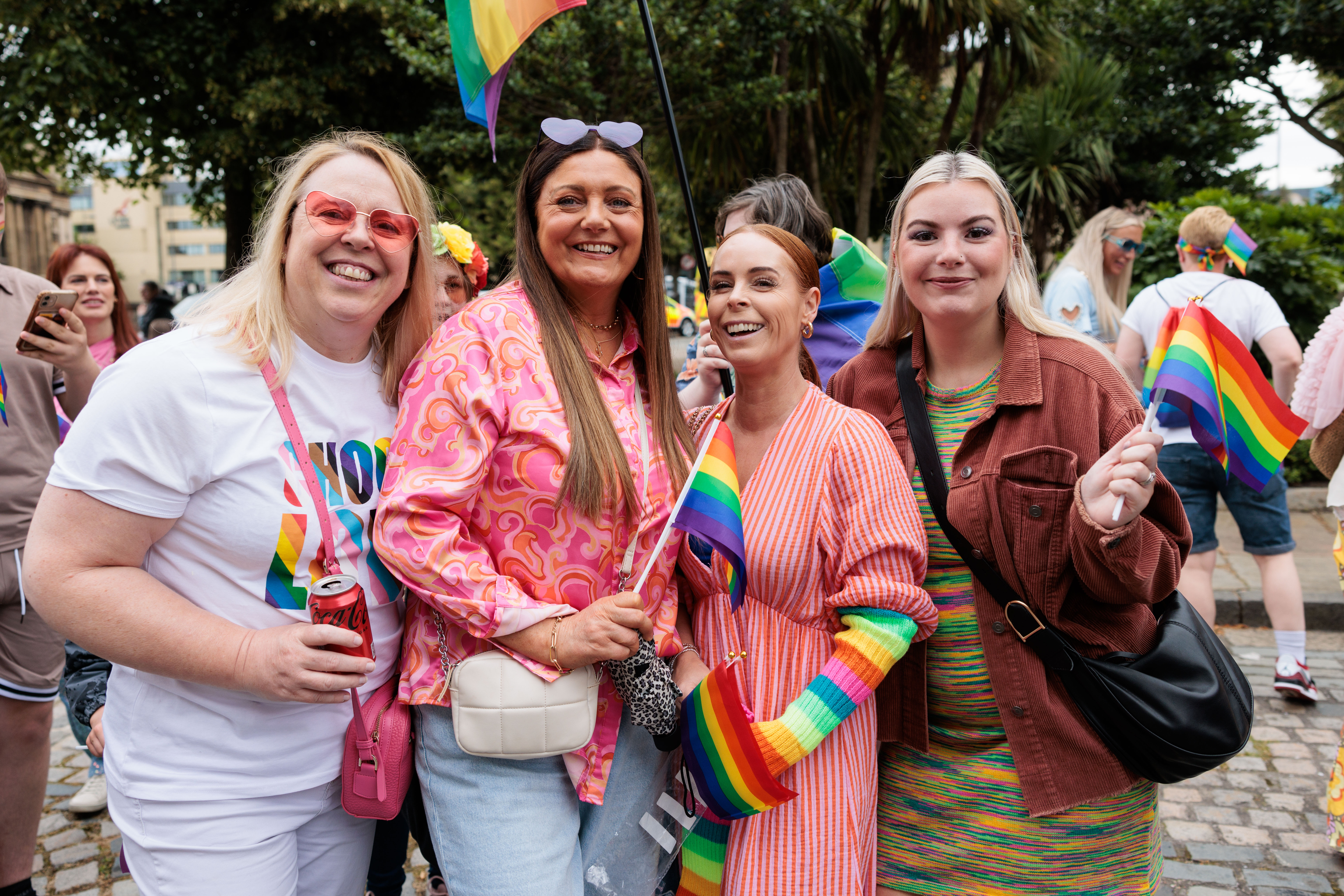 Four people standing together at an outdoor Pride event, holding rainbow flags. They are dressed in colourful clothing, with trees, other attendees, and Pride flags visible in the background.