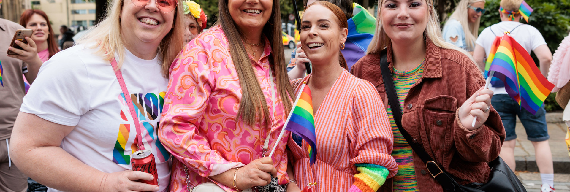 Four people standing together at an outdoor Pride event, holding rainbow flags. They are dressed in colourful clothing, with trees, other attendees, and Pride flags visible in the background.