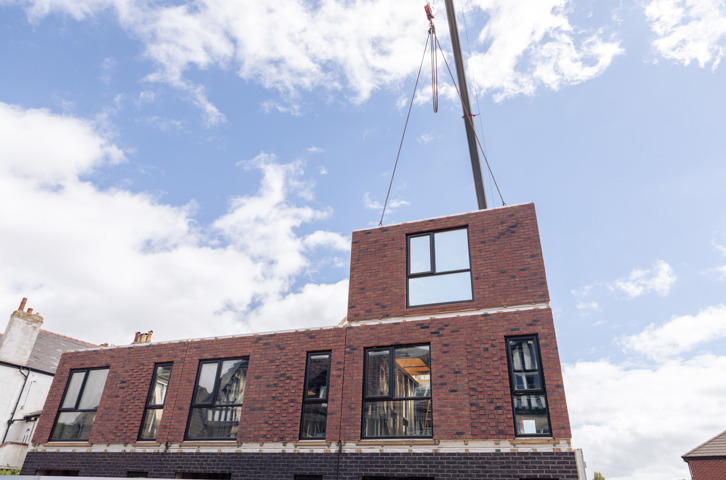 A crane lifts a brick wall section with a window into place on top of a new housing development, with a partly cloudy, blue sky in the background.