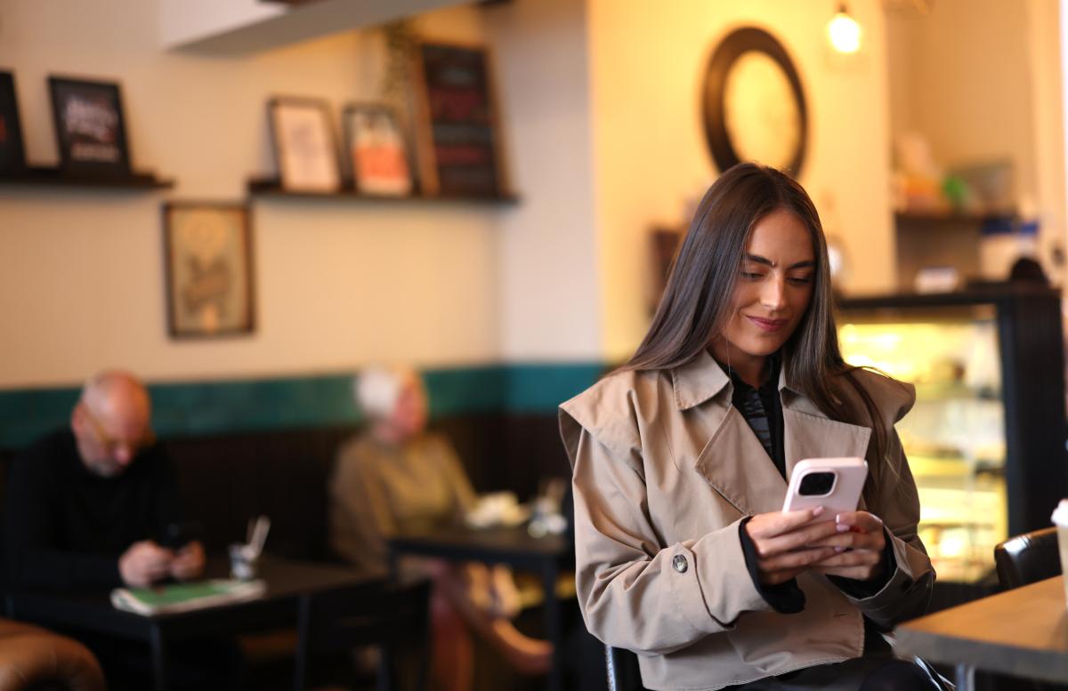 A person sitting in a café using a mobile phone, with other people seated in the background.