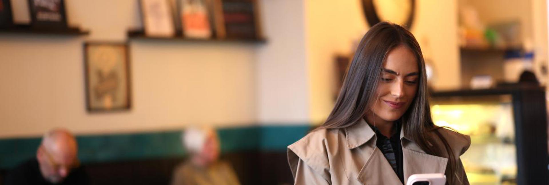 A person sitting in a café using a mobile phone, with other people seated in the background.