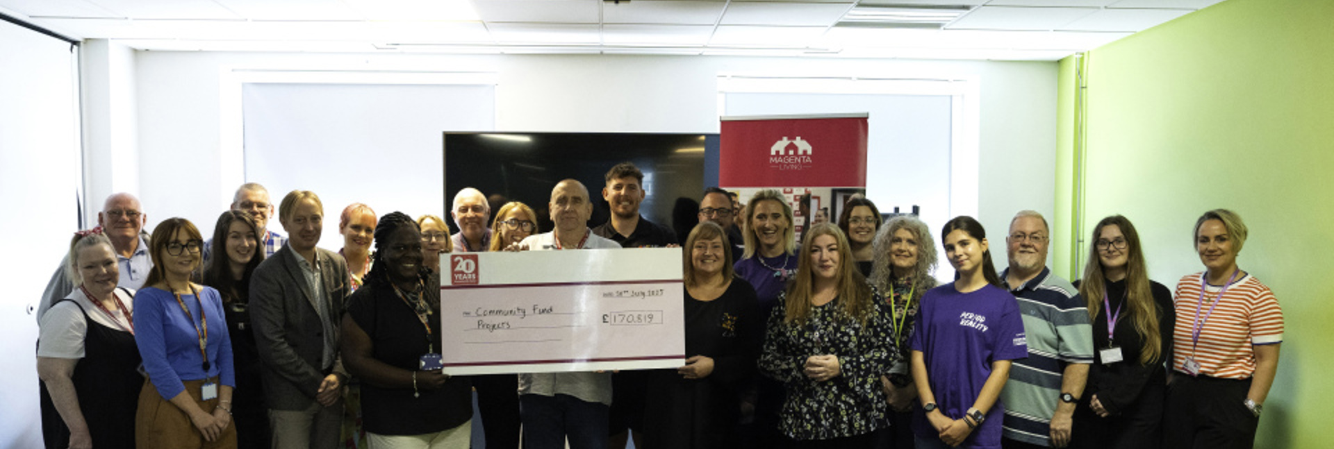 A large group of people standing together indoors holding an oversized cheque for a community fund projects.